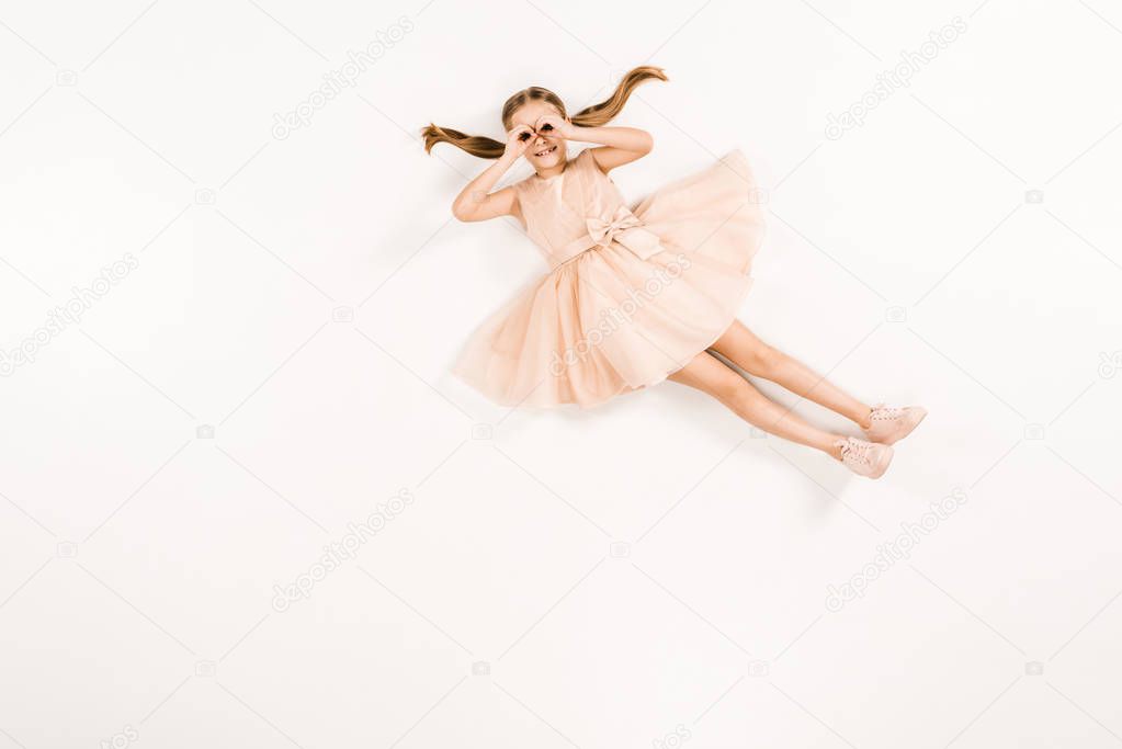 cheerful kid in dress with hands near eyes while lying on white 