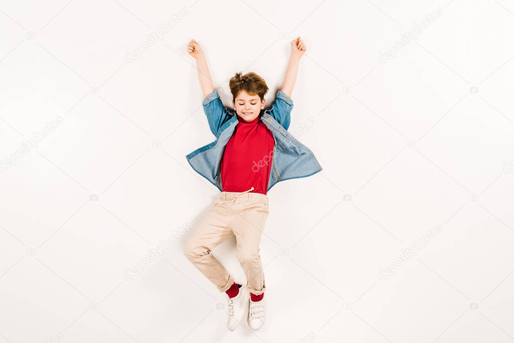 top view of cheerful kid with hands above head lying on white 