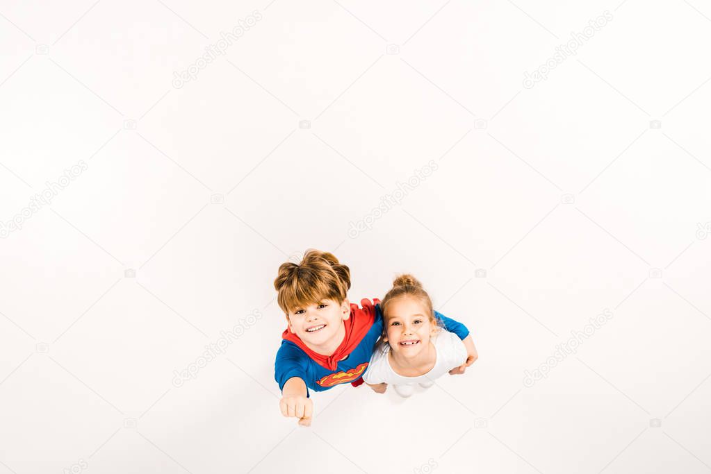 top view of happy kid in super hero costume hugging friend and gesturing on white 