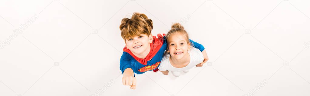 panoramic shot of happy kid in super hero costume hugging friend and gesturing on white 