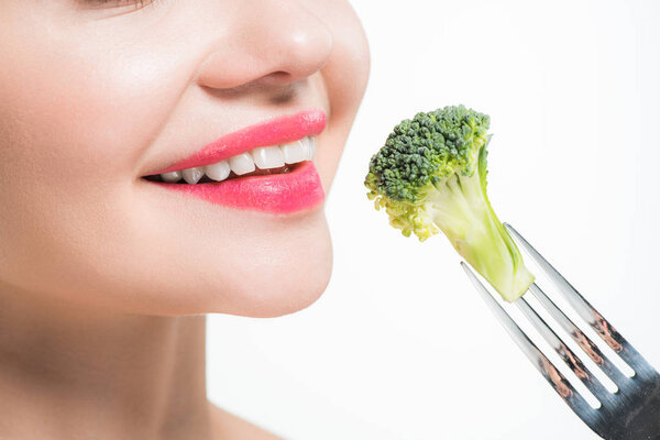 cropped view of happy woman holding silver fork with broccoli near pink lips isolated on white  