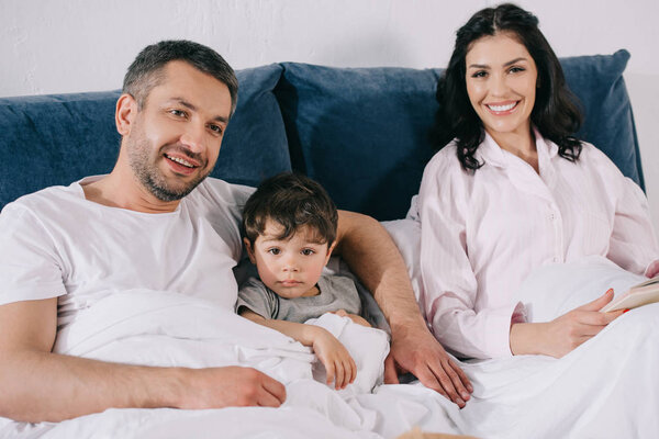 happy father smiling near toddler son and attractive wife in bedroom 