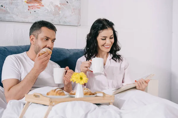 Wanita Bahagia Membaca Buku Dekat Suami Makan Croissant Tempat Tidur — Stok Foto