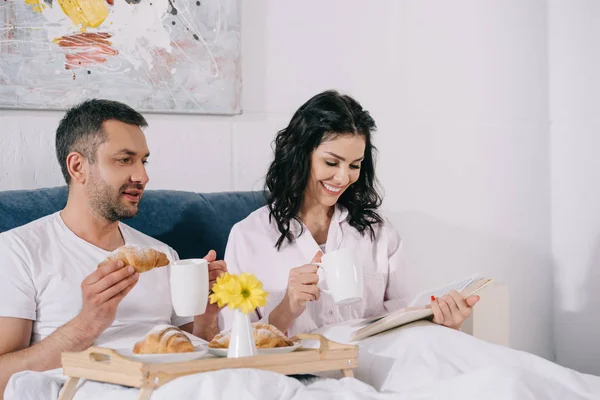 Happy Woman Reading Book Husband Holding Croissant Bed — Stock Photo, Image