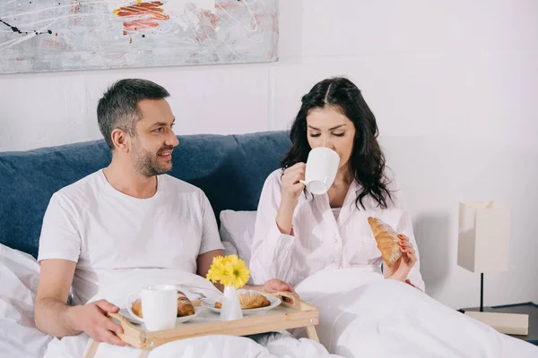 快乐的男人看着有吸引力的女人喝咖啡 并在床上拿着羊角面包 — 图库照片