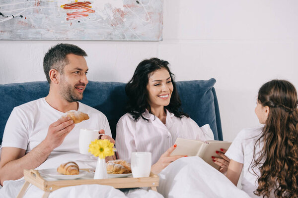 happy woman holding book near husband with cup and looking at daughter 