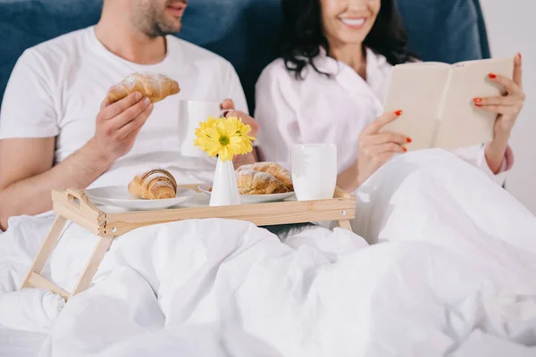Pemandangan Pria Ceria Memegang Croissant Dekat Wanita Membaca Tempat Tidur — Stok Foto