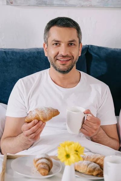 Εύθυμος Άνθρωπος Κρατώντας Γλυκό Κρουασάν Και Φλιτζάνι Καφέ Στο Κρεβάτι — Φωτογραφία Αρχείου
