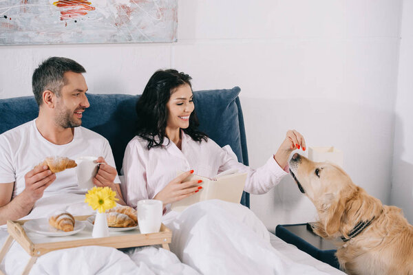 cheerful woman touching cute dog near man holding cup and croissant in bed 