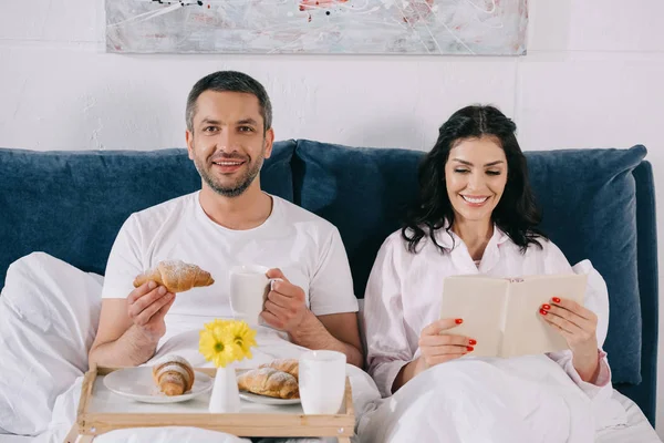 Feliz Mujer Leyendo Libro Cerca Alegre Marido Sosteniendo Croissant Taza — Foto de Stock