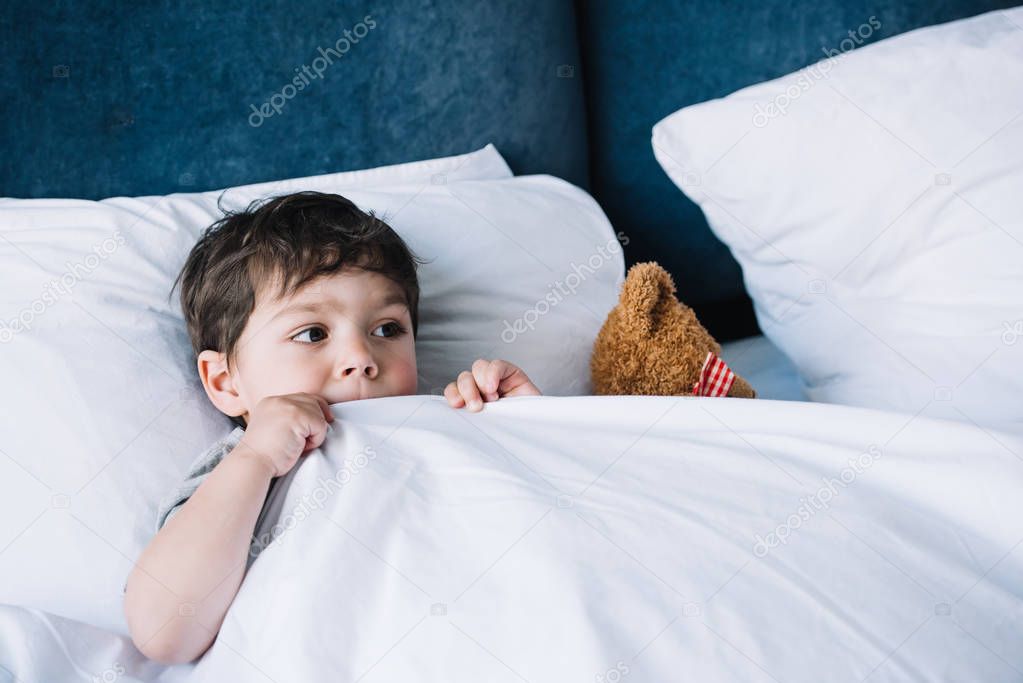 kid lying on white pillow in bed near teddy bear at home