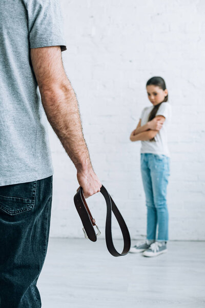 cropped view of father holding belt and upset daughter at home