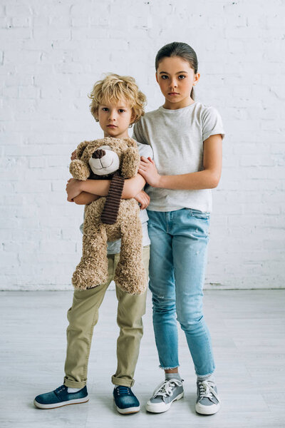 full length view of sad sister and brother with teddy bear at home