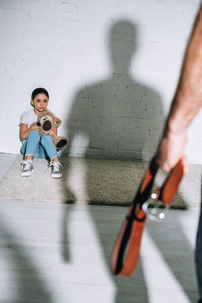 cropped view of father holding belt and scared daughter sitting on carpet near his silhouette