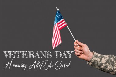 cropped view of soldier holding american flag near veterans day lettering on grey  clipart