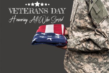 cropped view of man in military uniform holding flag of america near veterans day lettering on grey  clipart