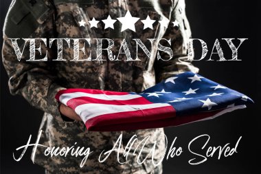 cropped view of soldier in military uniform holding flag of america near veterans day lettering on grey  clipart