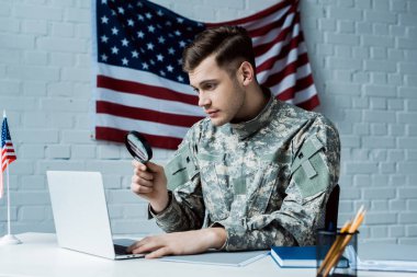 young soldier in uniform holding magnifying glass near laptop in office 