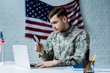 young soldier in uniform holding magnifying glass near laptop in office 