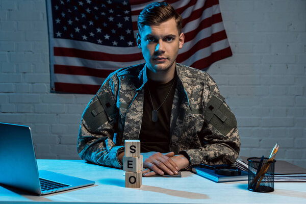 handsome military man sitting with clenched hands near laptop and cubes with seo lettering 