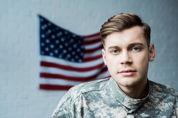 handsome man in military uniform looking at camera near american flag 