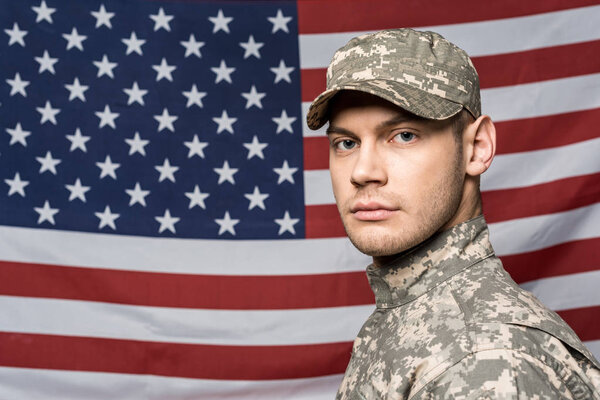 handsome man in military uniform and cap looking at camera near flag of america
