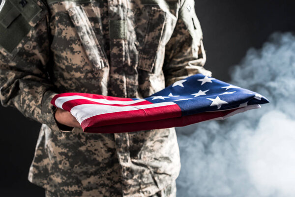 cropped view of man in military uniform holding flag of america on black with smoke 