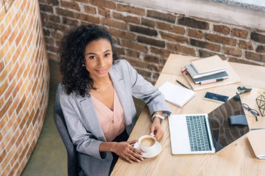 ofiste masada kahve fincanı ve dizüstü bilgisayar ile gülümseyen afrikalı amerikalı Casual iş kadını