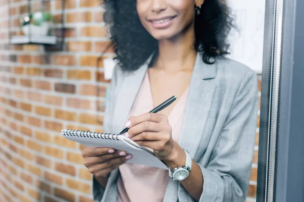 cropped view of smiling african american Casual businesswoman writing in notebook in loft office