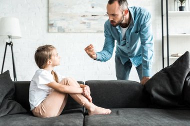 angry father threatening with fist and standing near upset daughter at home  clipart