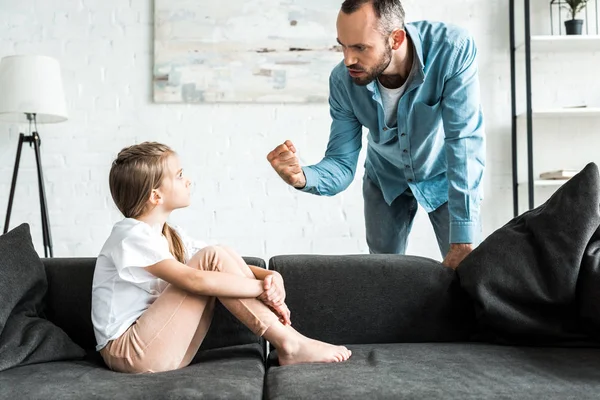 angry father threatening with fist and standing near upset daughter at home