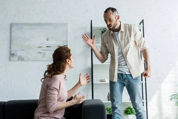 Homem Zangado Gesticulando Enquanto Olha Para Esposa Sentada Sofá Casa — Fotografia de Stock