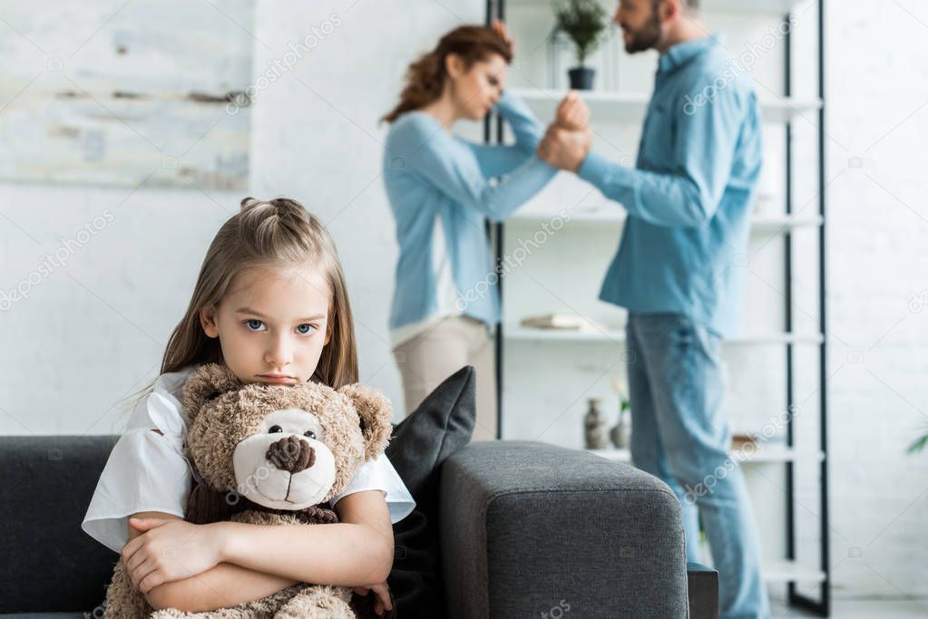 selective focus of sad kid holding teddy bear near quarreling parents at home 