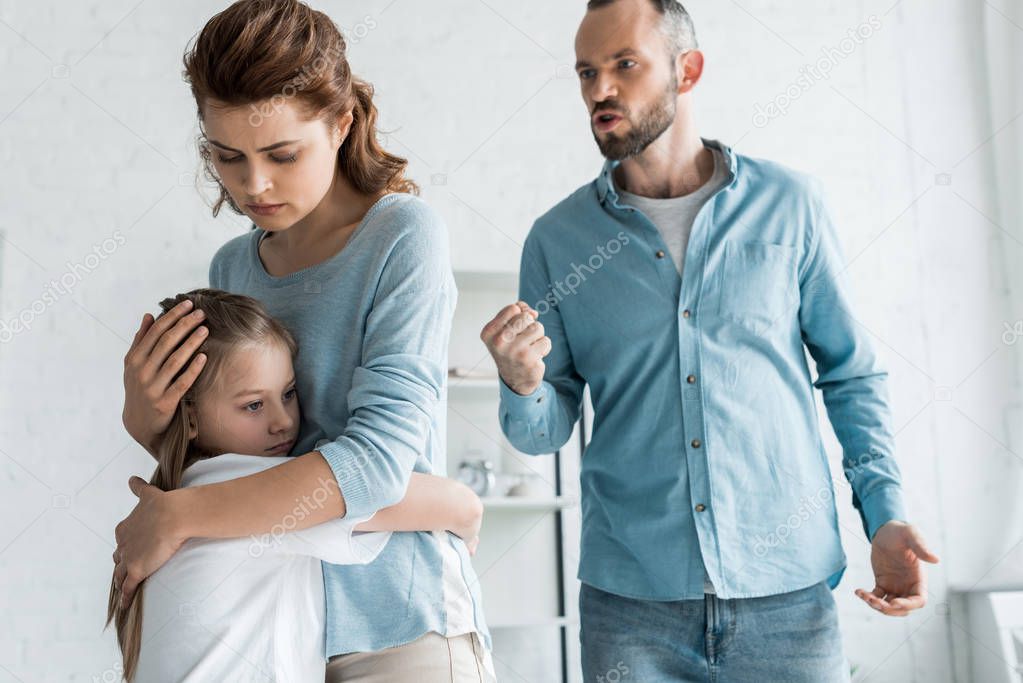 upset mother hugging daughter while angry man showing fist at home 