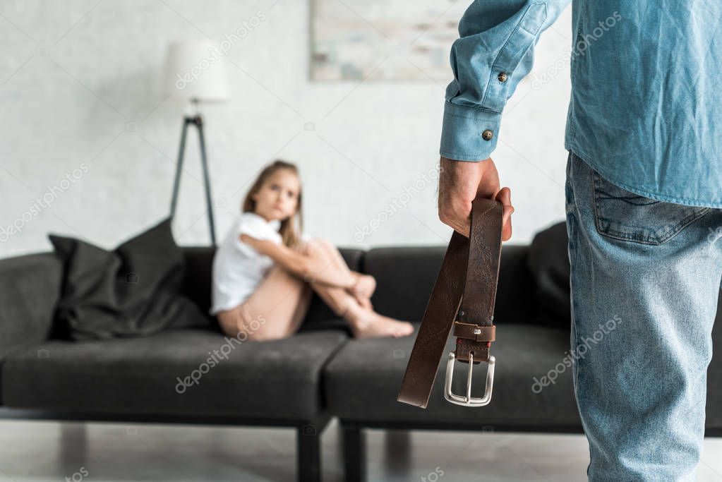 cropped view of father holding belt near scared daughter sitting on sofa 