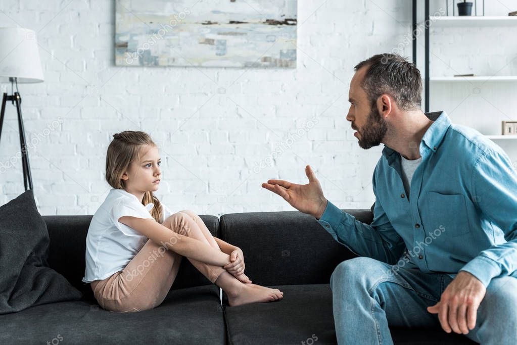 angry father gesturing near upset kid sitting on sofa 