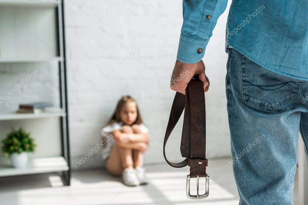 selective focus of angry man holding belt and standing near scared kid sitting on floor 