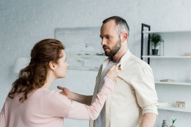 angry woman touching shirt of handsome bearded man at home  clipart