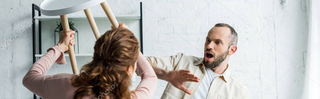 panoramic shot of surprised man looking at angry holding chair while threatening at home 