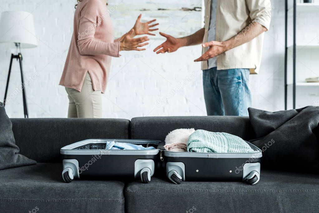 cropped view of man and woman standing and gesturing near suitcase with clothes on sofa 