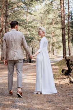 full length view of smiling just married couple holding hands and walking in forest clipart