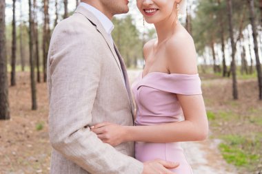 cropped view of happy just married couple embracing in forest with smile clipart