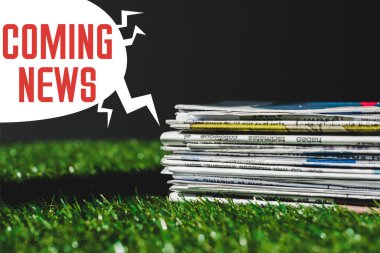 stack of different print newspapers on fresh green grass near speech bubble with red coming news lettering isolated on black clipart