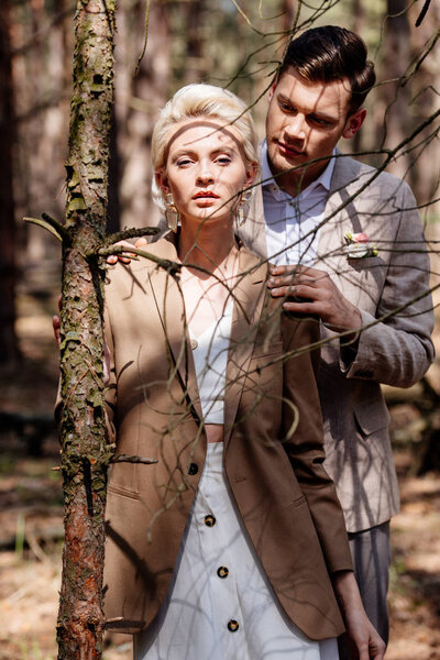 stylish man gently embracing blonde bride in forest