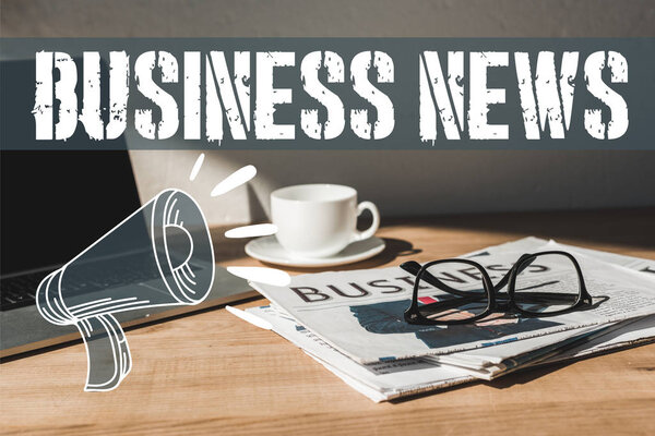 стаканы на деловую газету возле ноутбука и чашки с блюдцем на деревянном столе с бизнес-новостями и иллюстрацией громкоговорителя
