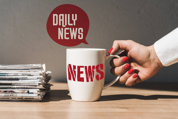 cropped view of woman holding white cup with news lettering on wooden table near pile of different newspapers and speech bubble illustration with daily news text