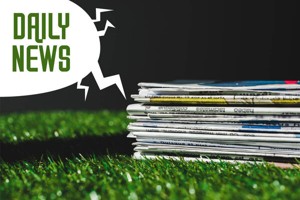 stack of different print newspapers on fresh green grass near speech bubble with green daily news lettering isolated on black