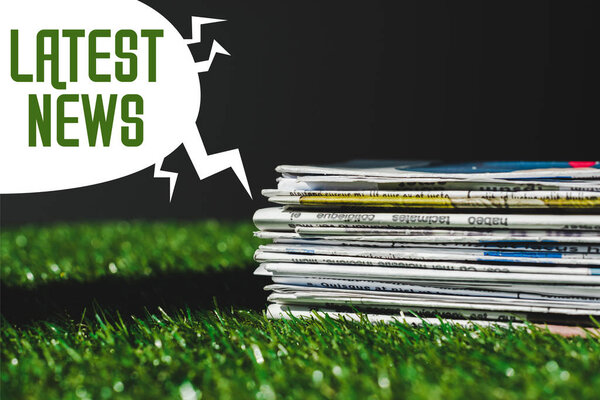 stack of different print newspapers on fresh green grass near speech bubble with green latest news lettering isolated on black