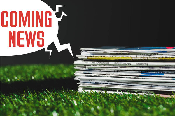 stack of different print newspapers on fresh green grass near speech bubble with red coming news lettering isolated on black