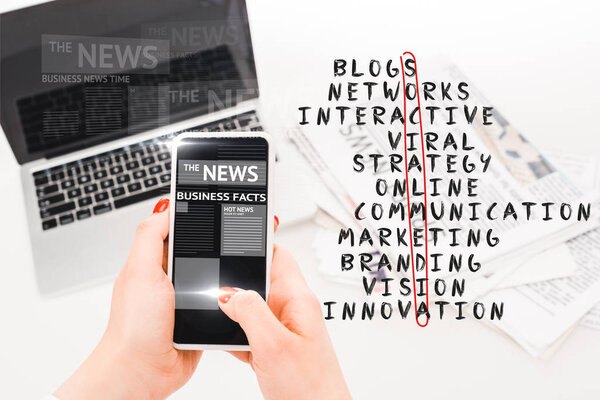 selective focus of woman holding smartphone with business news illustration near laptop, newspapers and social media crossword